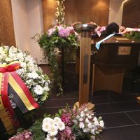 Un cercueil de bois brun encerclé de nombreuses fleurs rouges et rose et recouvert de petits bouquets portant des rubans roses au nom de ses enfants, et petites-filles.